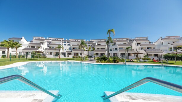 A swimming pool with a clear blue water, surrounded by a row of white buildings with balconies, and palm trees.