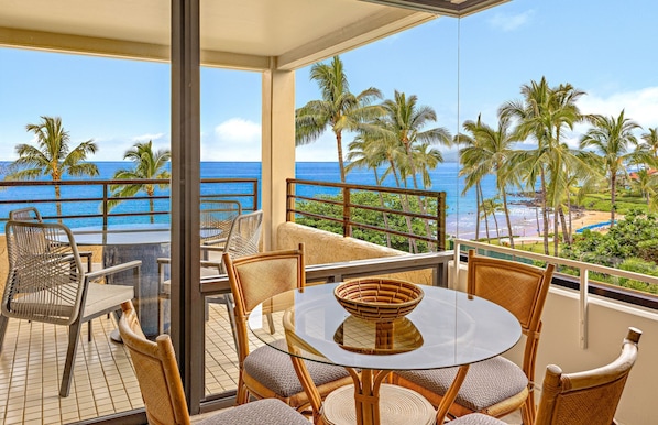 A balcony with wicker furniture, a glass table, and a view of the ocean and palm trees.