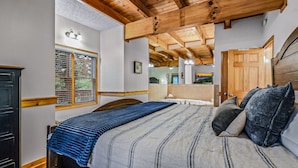 A bedroom with a wooden ceiling, a large bed with a striped bedspread, a window with blinds, a wooden wardrobe, and a wooden door.