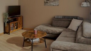 A living room with a sectional sofa, a round coffee table, a TV on a wooden cabinet, and a pendant light. - Cosy apartment for 4 people with WIFI, TV and balcony (Fuhre)