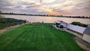 Une pelouse verdoyante agrémentée d’un belvédère et d’une petite construction, dominant une étendue d’eau sur laquelle se reflète un coucher de soleil.