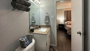 A bathroom with a sink, mirror, and towel rack. There is a bed visible in the adjacent room.