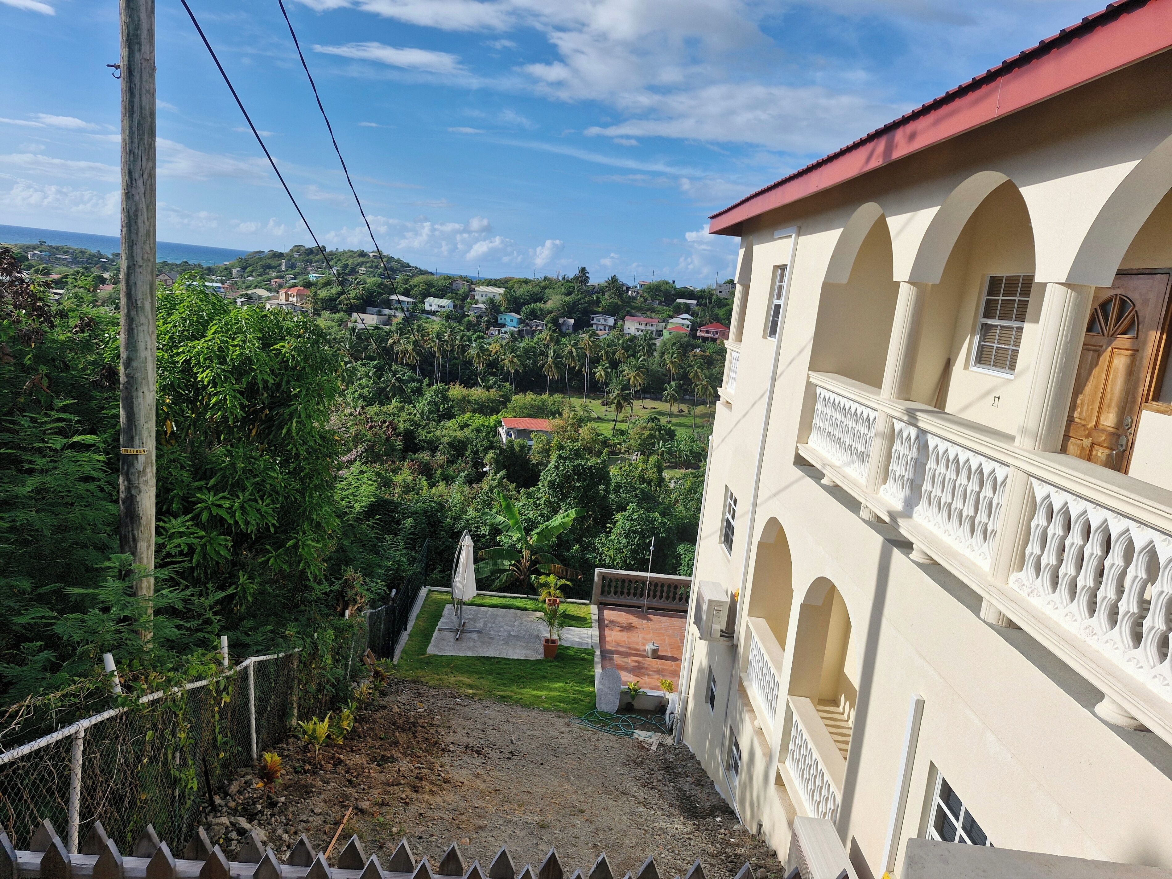 A view from a balcony overlooking a lush green landscape with a building featuring arched balconies and a red roof.