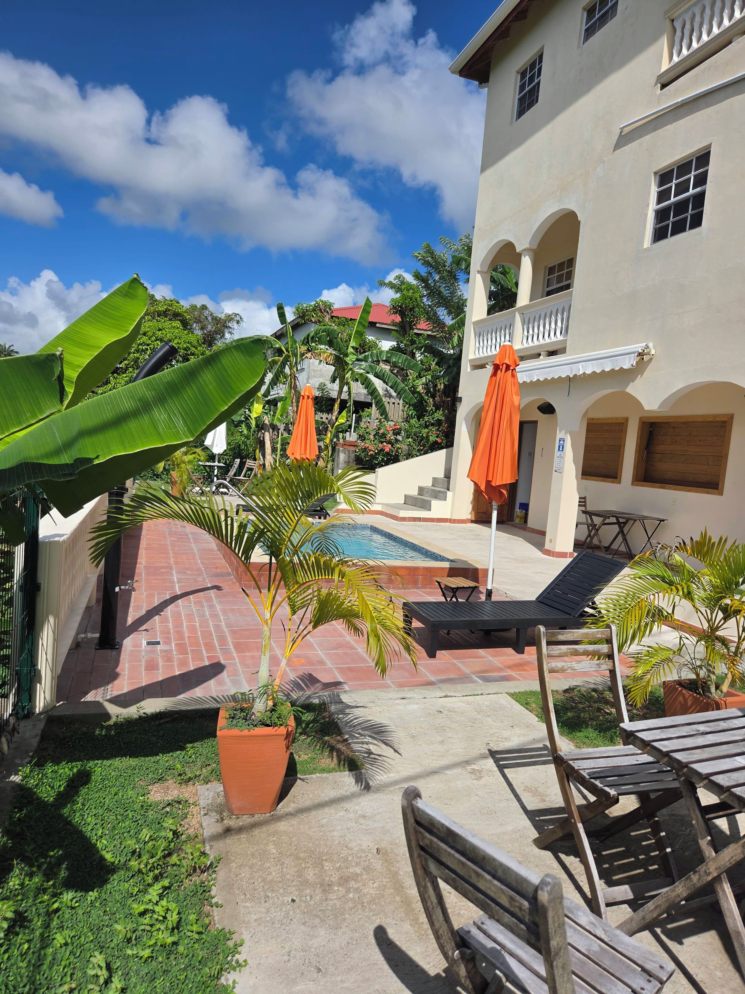 Área de piscina com espreguiçadeiras e guarda-sóis, cercada por plantas em vasos e um edifício com arcos.
