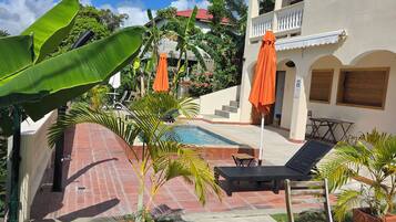 Área de piscina com espreguiçadeiras e guarda-sóis, cercada por plantas em vasos e um edifício com arcos.