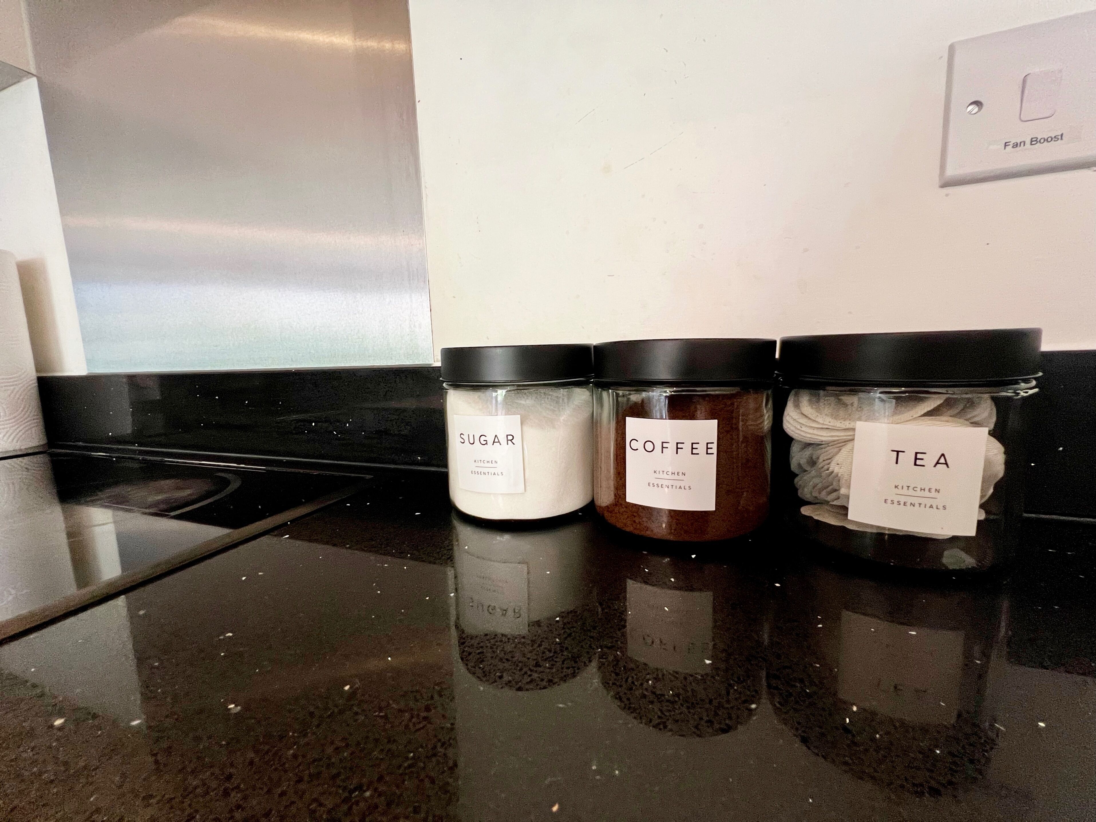 Three kitchen essentials jars labeled 'SUGAR,' 'COFFEE,' and 'TEA' on a kitchen counter.
