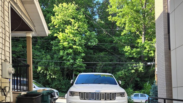 Un Jeep blanco estacionado en una entrada de casa, con una vivienda a un lado y una calle arbolada al otro.