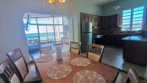 Un coin repas avec une table dressée pour quatre personnes, une cuisine équipée d’appareils modernes et une vue sur l’océan grâce à de grandes fenêtres.