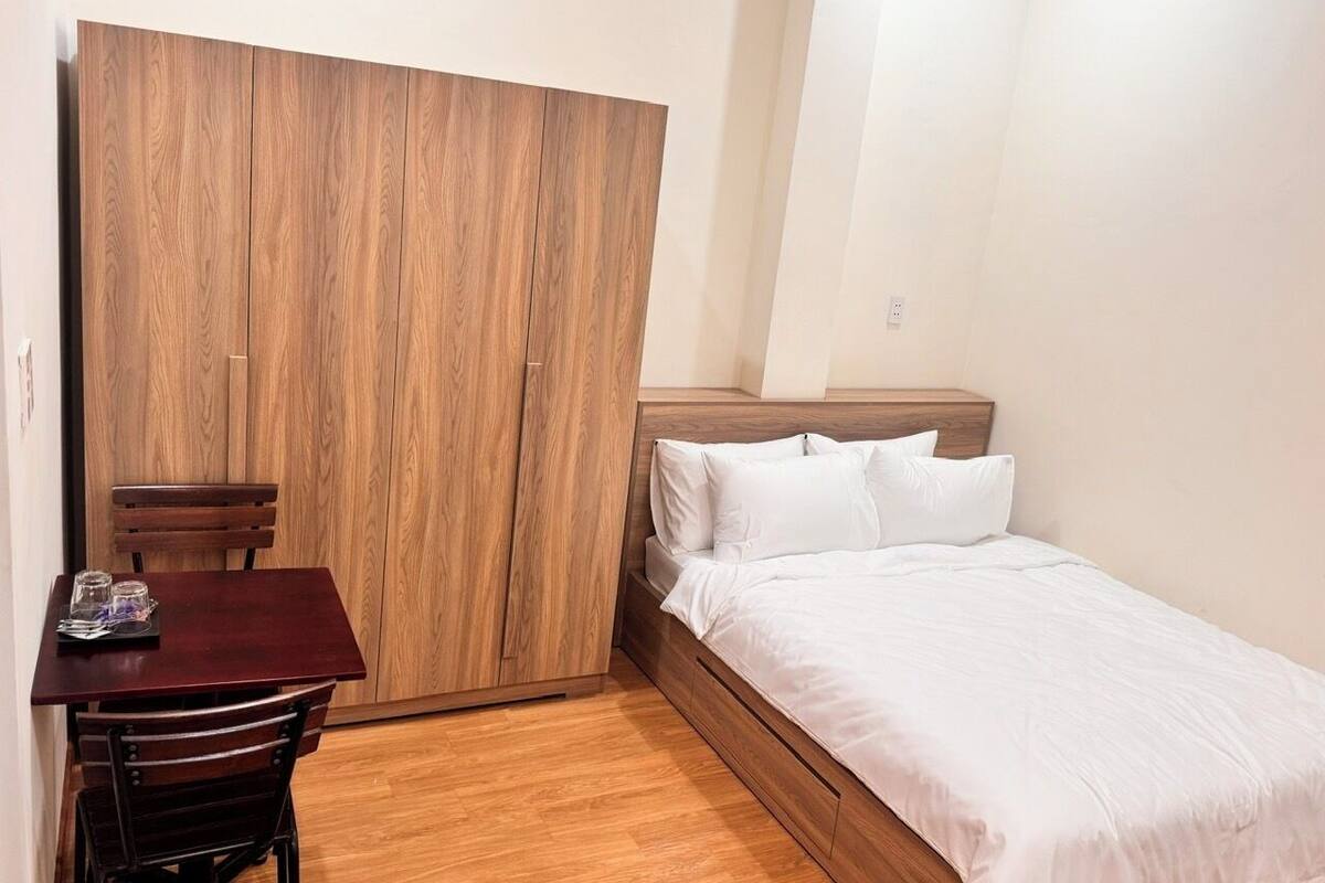A bedroom with a wooden wardrobe, a bed with white bedding, a small table with chairs, and a wooden floor.
