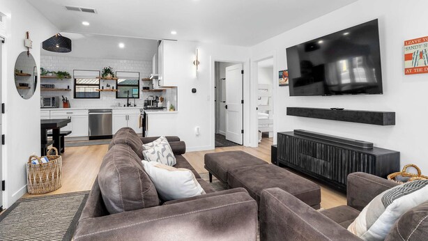 Una sala moderna con un sofá gris, un televisor de pantalla plana montado en la pared y una cocina visible a través de una puerta abierta.