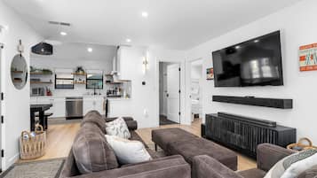 A modern living room with a grey sofa set, a flat-screen TV mounted on the wall, and a kitchen visible through an open door.