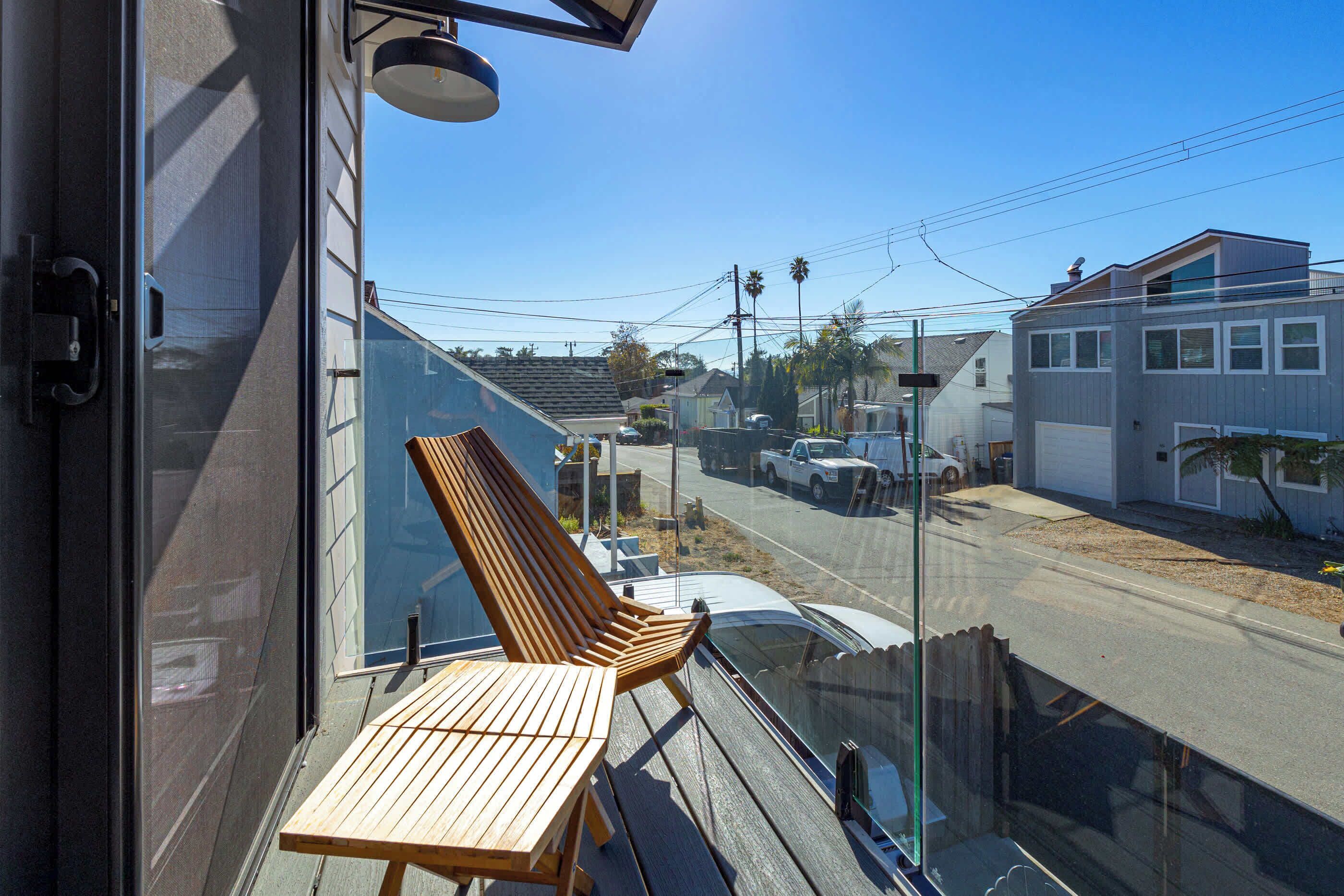 A balcony with a wooden chair and table, overlooking a street with houses and parked cars.