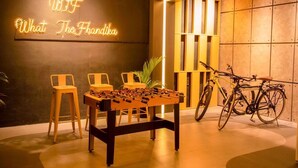 Una mesa de fútbol de mesa, dos sillas, un letrero de neón y una bicicleta en una habitación moderna con pared oscura y detalles de madera.