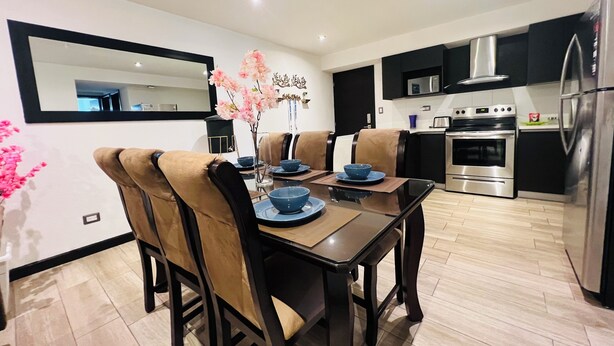 A modern kitchen with a dining area, featuring a large mirror, black cabinets, stainless steel appliances, and a vase with pink flowers on the table.