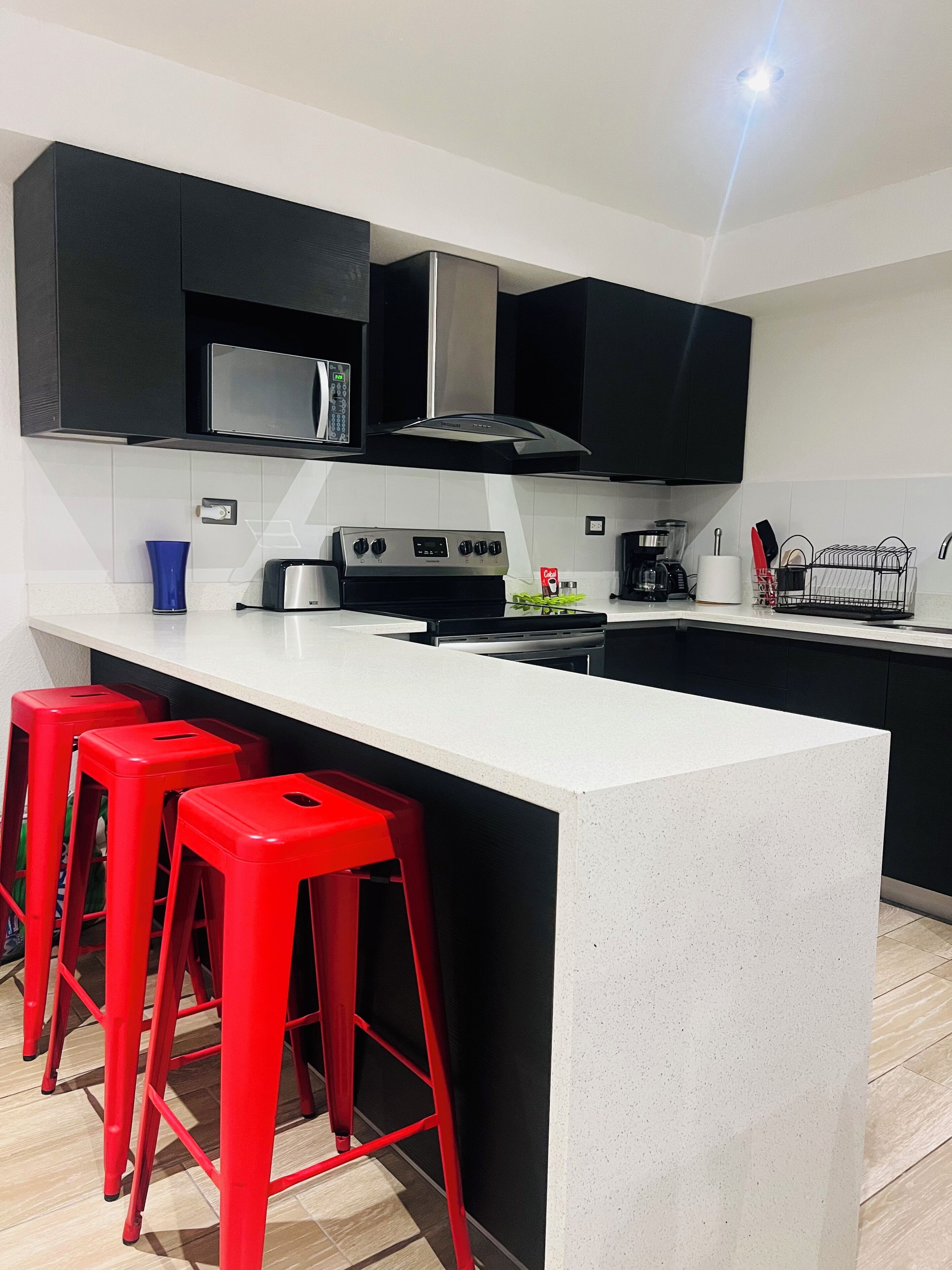 A modern kitchen with black cabinets, a white countertop, a stainless steel stove, and red bar stools.