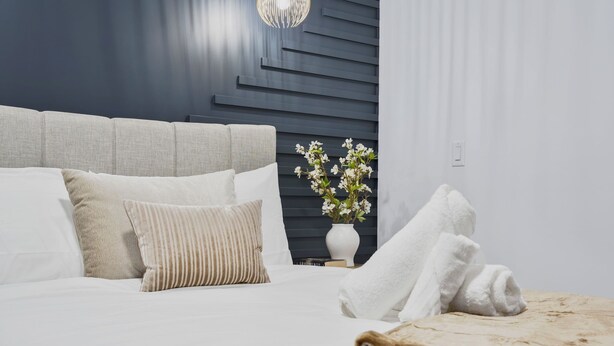 Un lit bien fait, avec du linge de lit blanc et un plaid beige, un vase avec des fleurs blanches et un motif mural moderne.