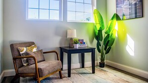 A room with a chair, a small table with a lamp, a potted plant, and two windows. - We Connect Co-Living (St. Louis Park)