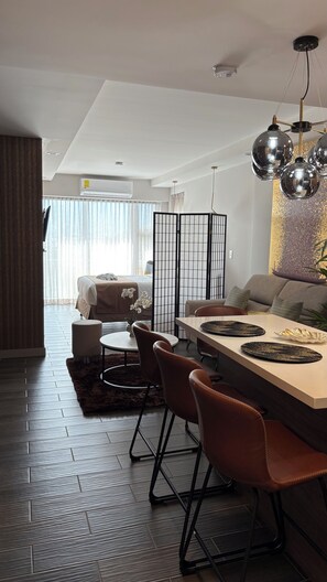 A modern hotel room with a dining area featuring a large table, four chairs, and a decorative chandelier.