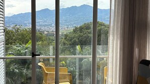A balcony with a view of a cityscape and mountains. There is a wicker chair and a glass railing. - Cozy Apartment 10 min to Airport Be Cariari Condo new (Ulloa)
