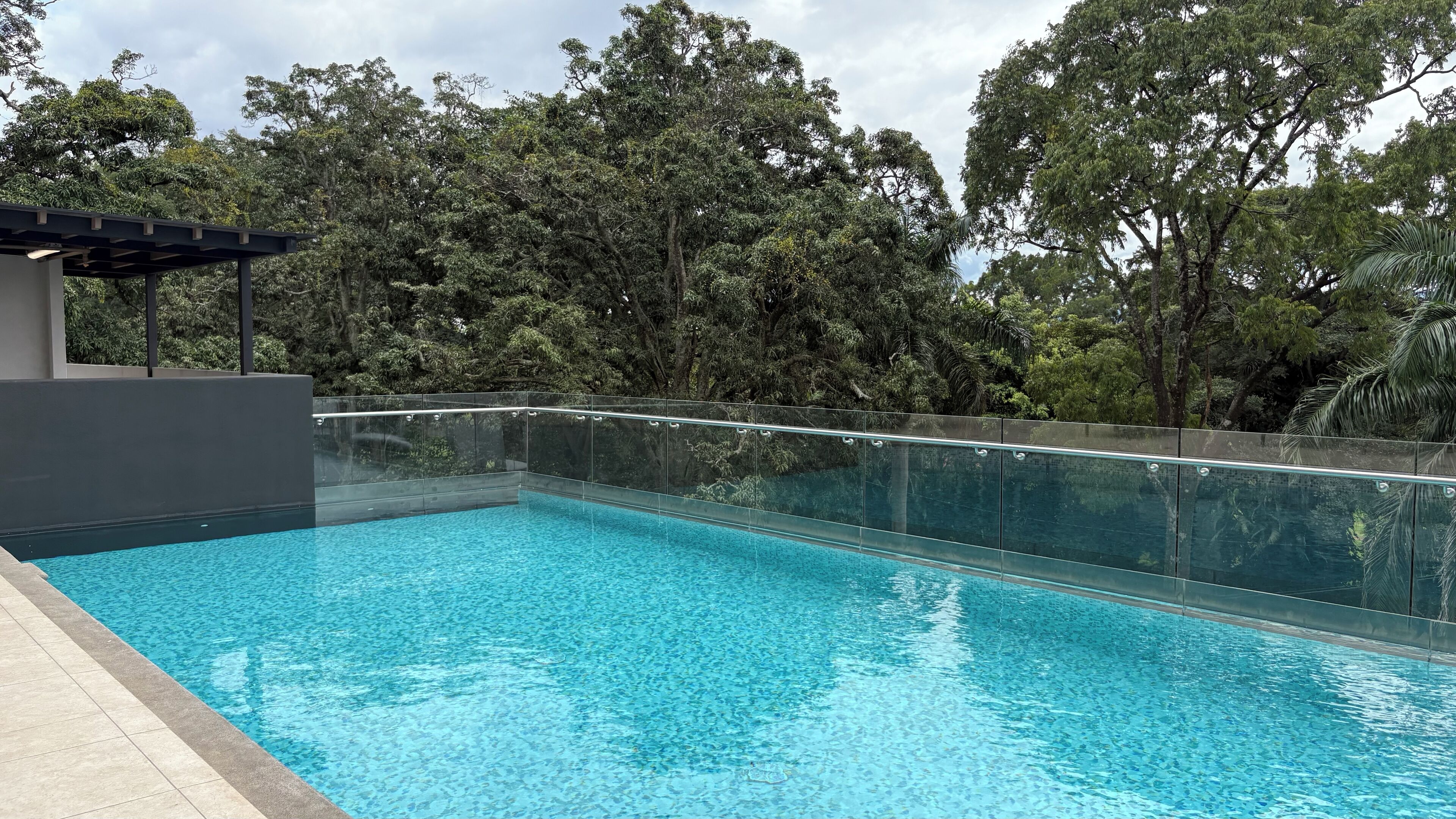 Una piscina rectangular con agua azul cristalina, rodeada por una barandilla de vidrio y una estructura de edificio moderna.