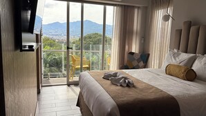 A hotel room with a bed, a TV mounted on the wall, a balcony with a chair, and a view of mountains. - Cozy Apartment 10 min to Airport Be Cariari Condo new (Ulloa)