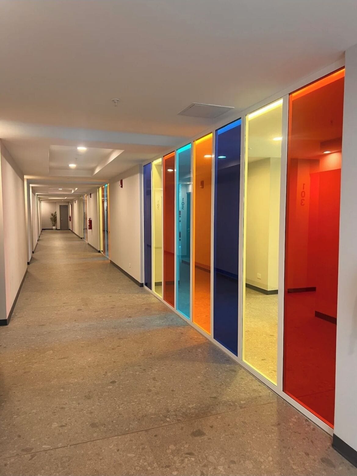 Corredor extenso com divisórias de vidro coloridas e piso de concreto.