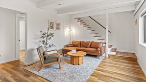 Un salon moderne avec un plancher en bois, un canapé en cuir marron, une table basse en bois et une chaise en bois. On y trouve un escalier avec des marches en bois et un mur blanc orné d’un petit objet décoratif.