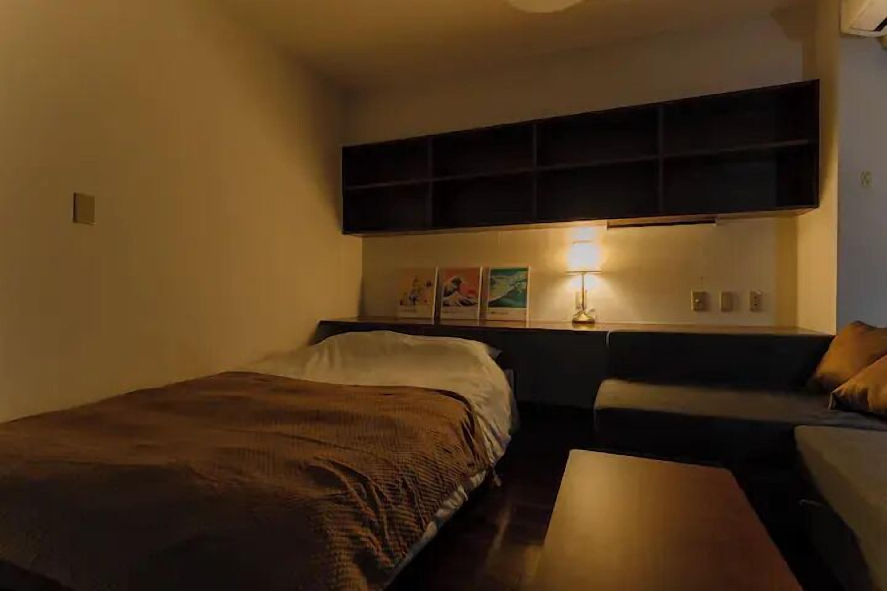 A small, dimly lit room with a single bed, a sofa, and a small table. There are wall-mounted shelves above the sofa and a wall-mounted light fixture.