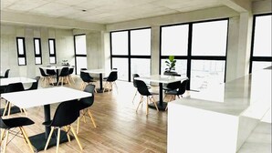 Un moderno salón de conferencias con varias mesas y sillas, amplias ventanas con vistas a la ciudad y suelo de madera.