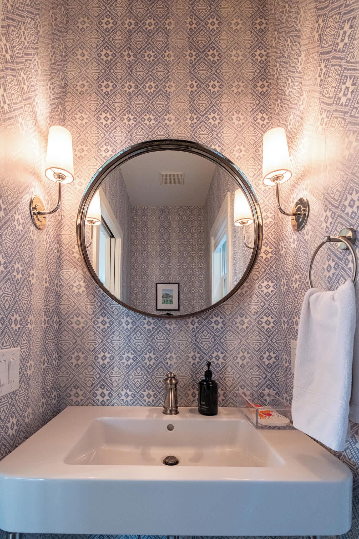 A bathroom with a round mirror, two wall-mounted lights, a sink, and patterned wallpaper.