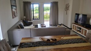 Un salon avec un canapé, une table à manger surmontée d’un panier en osier, un téléviseur et un balcon agrémenté de rideaux.