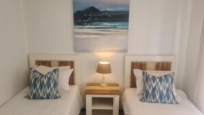 Une chambre avec deux lits, chacun agrémenté d’un coussin à motifs, une table de chevet avec une lampe et un tableau représentant un paysage marin au-dessus des lits.