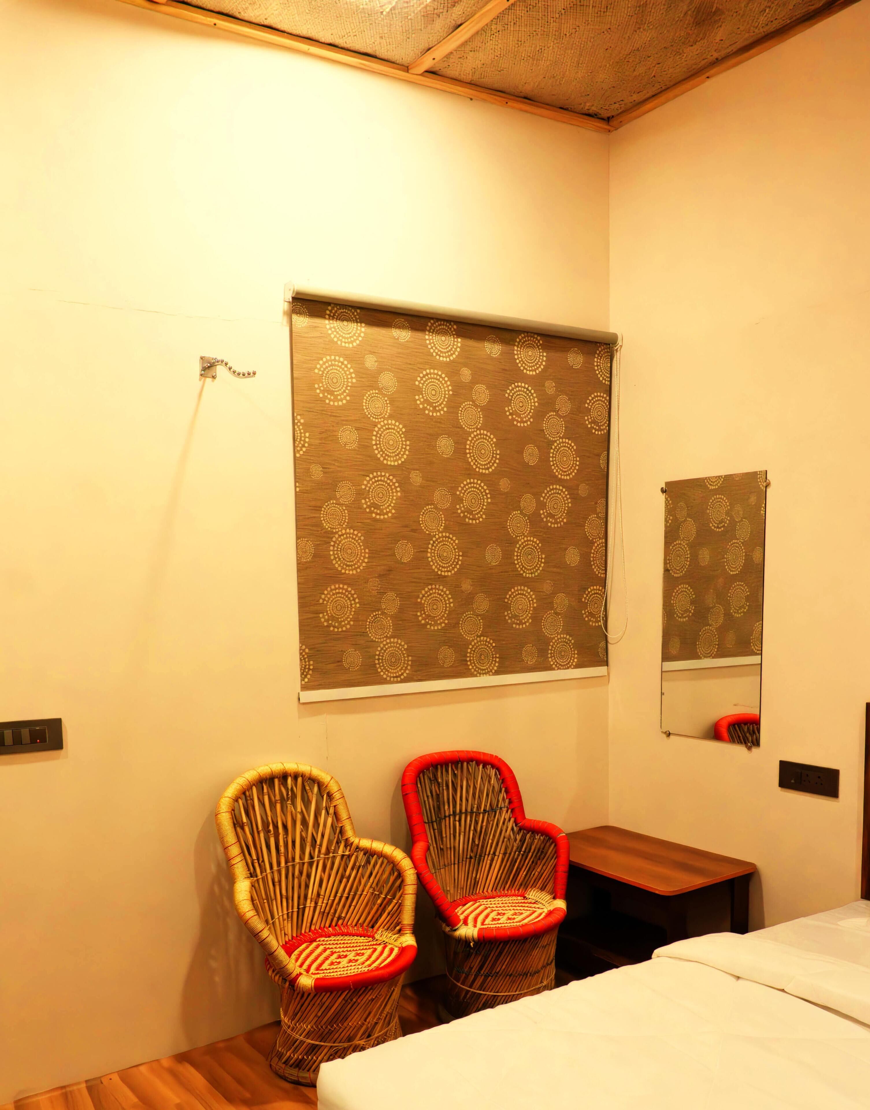 Una habitación con dos sillones de mimbre, uno con asiento rojo, una cama con ropa blanca, una mesita de noche de madera, un espejo adosado a la pared y una cortina estampada.