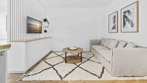 A modern living room with a sofa, a coffee table, and a large TV mounted on a white wall with vertical stripes. There are two framed pictures on the wall and a small plant on the shelf. - Studio3: Chic Bondi Abode with Pool & A/C (Bondi Junction)
