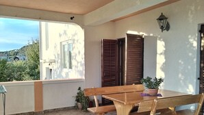 Un banco y una mesa de madera en un balcón con una planta en maceta, una ventana con contraventanas y una lámpara fijada a la pared.