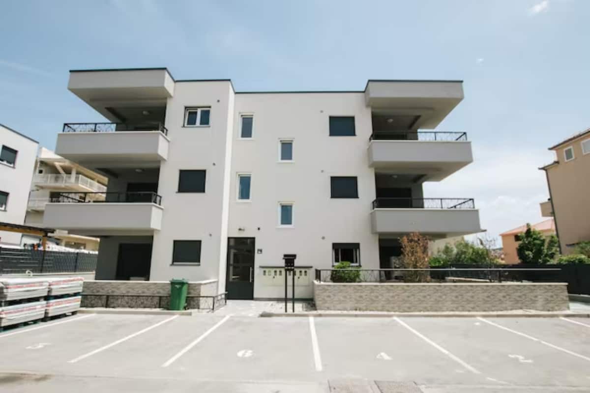 A modern multi-story building with balconies, a parking lot, and a small garden area.