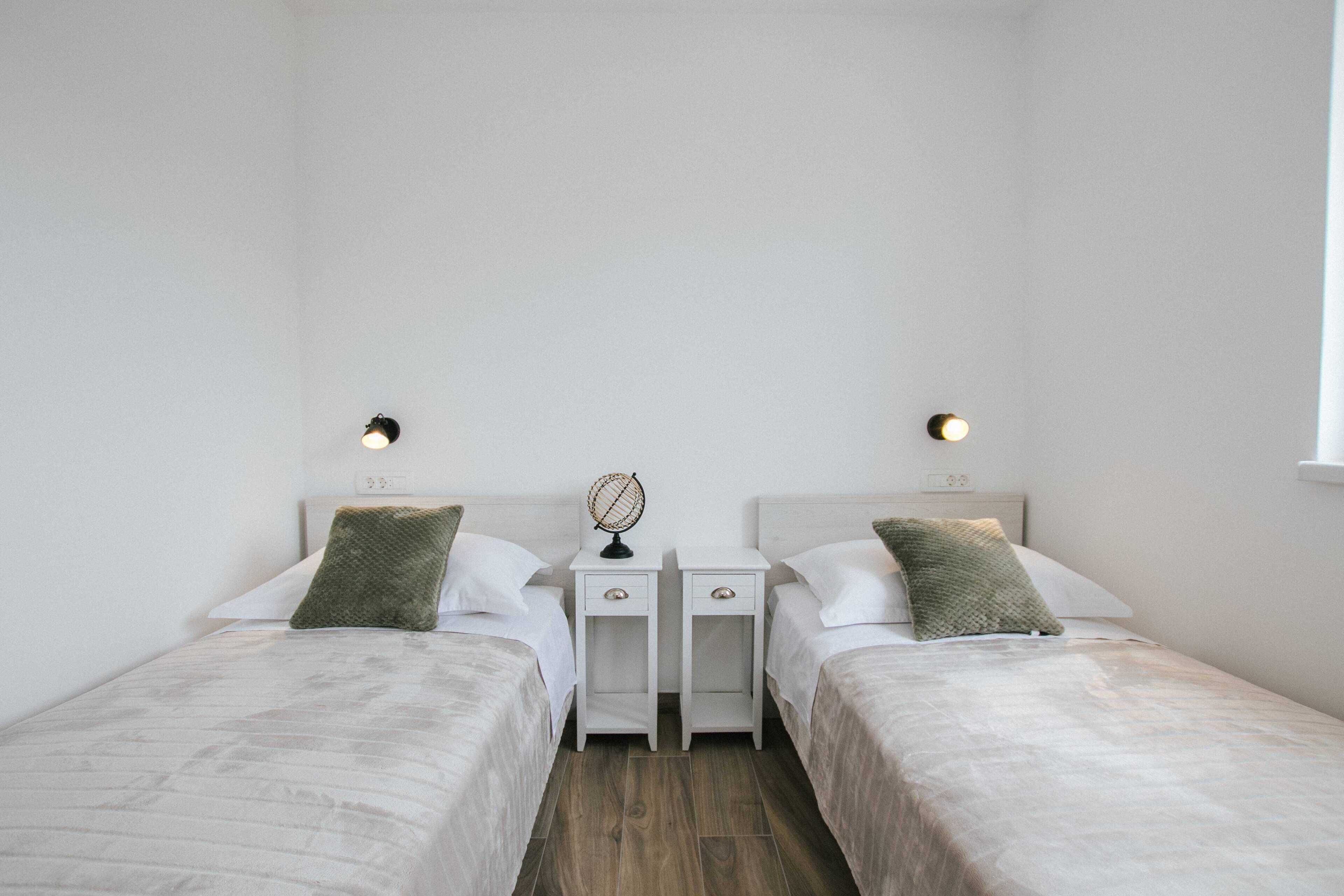 A bedroom with two beds, white bedding, a small table with a globe, and wall-mounted lights.