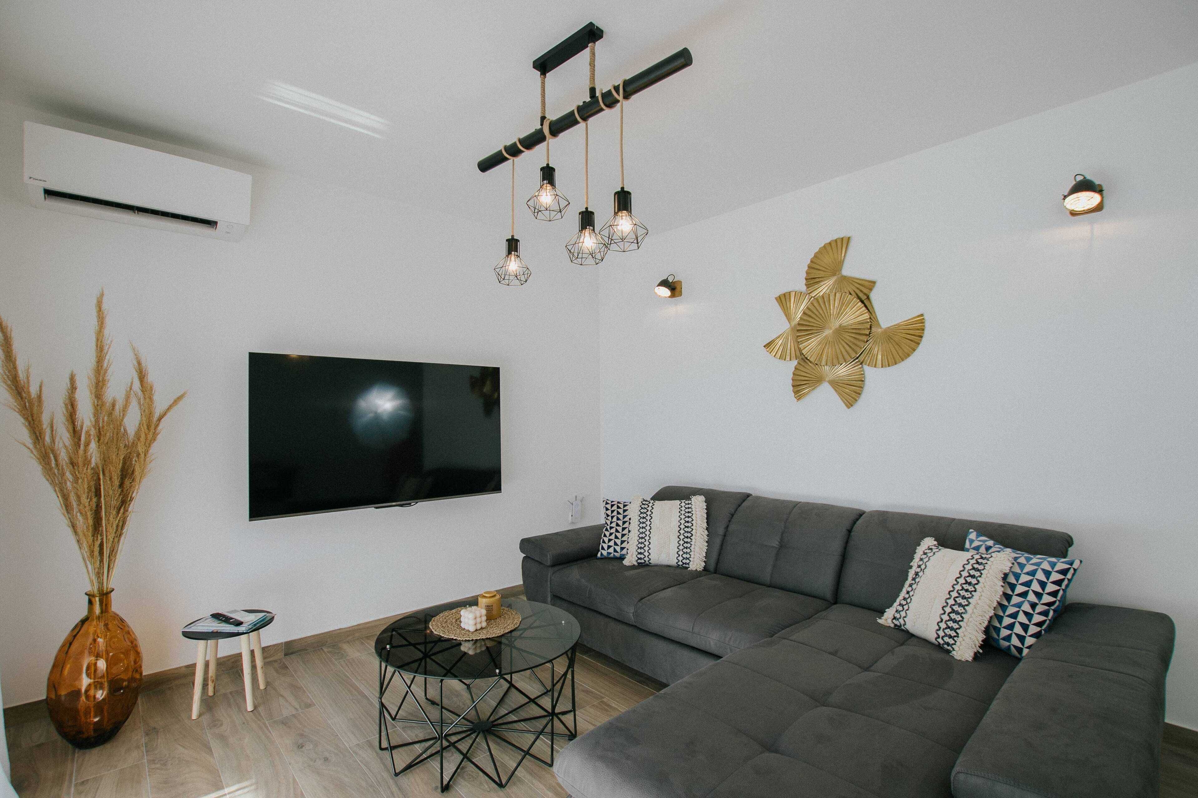 Un salon moderne avec un canapé d’angle gris, un téléviseur à écran plat, un élément décoratif mural et un luminaire suspendu original.