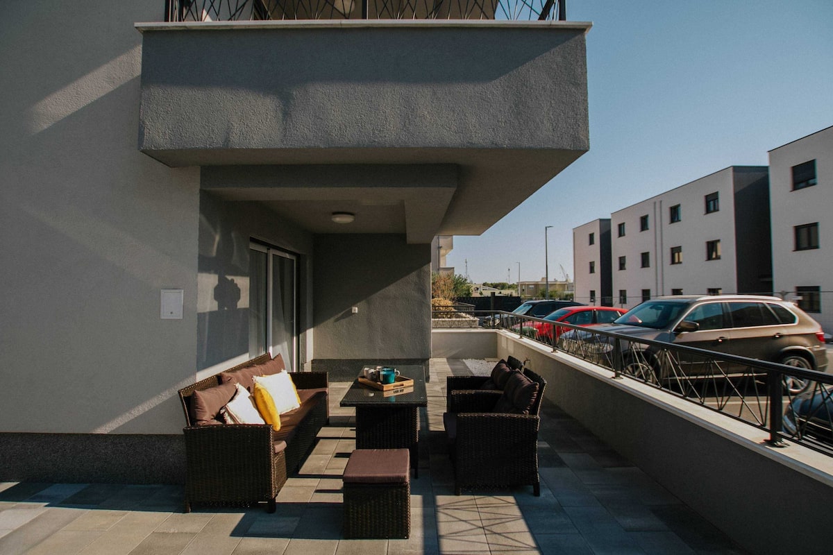 A balcony with wicker furniture, a table, and a view of parked cars and buildings.