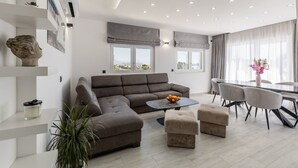 A modern living room with a sectional sofa, a coffee table, ottomans, a dining area with chairs, and a large window with a view. - Scenic Kastel Luksic Apartments (Kastela)