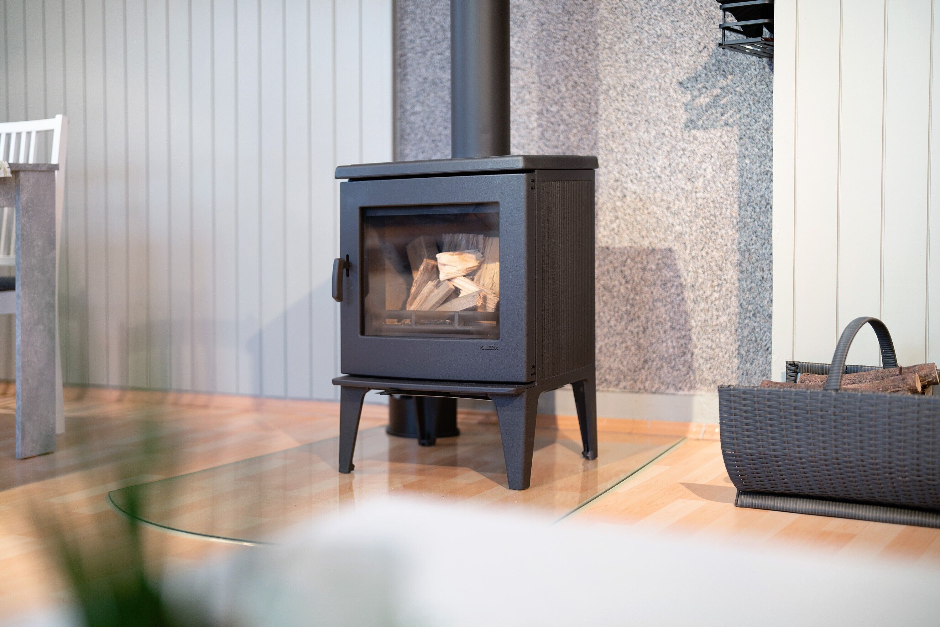 A modern wood-burning stove with a glass door, a metal basket, and a wooden floor.