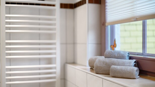 A bathroom with white tiled walls, a window with blinds, and towels on a shelf.