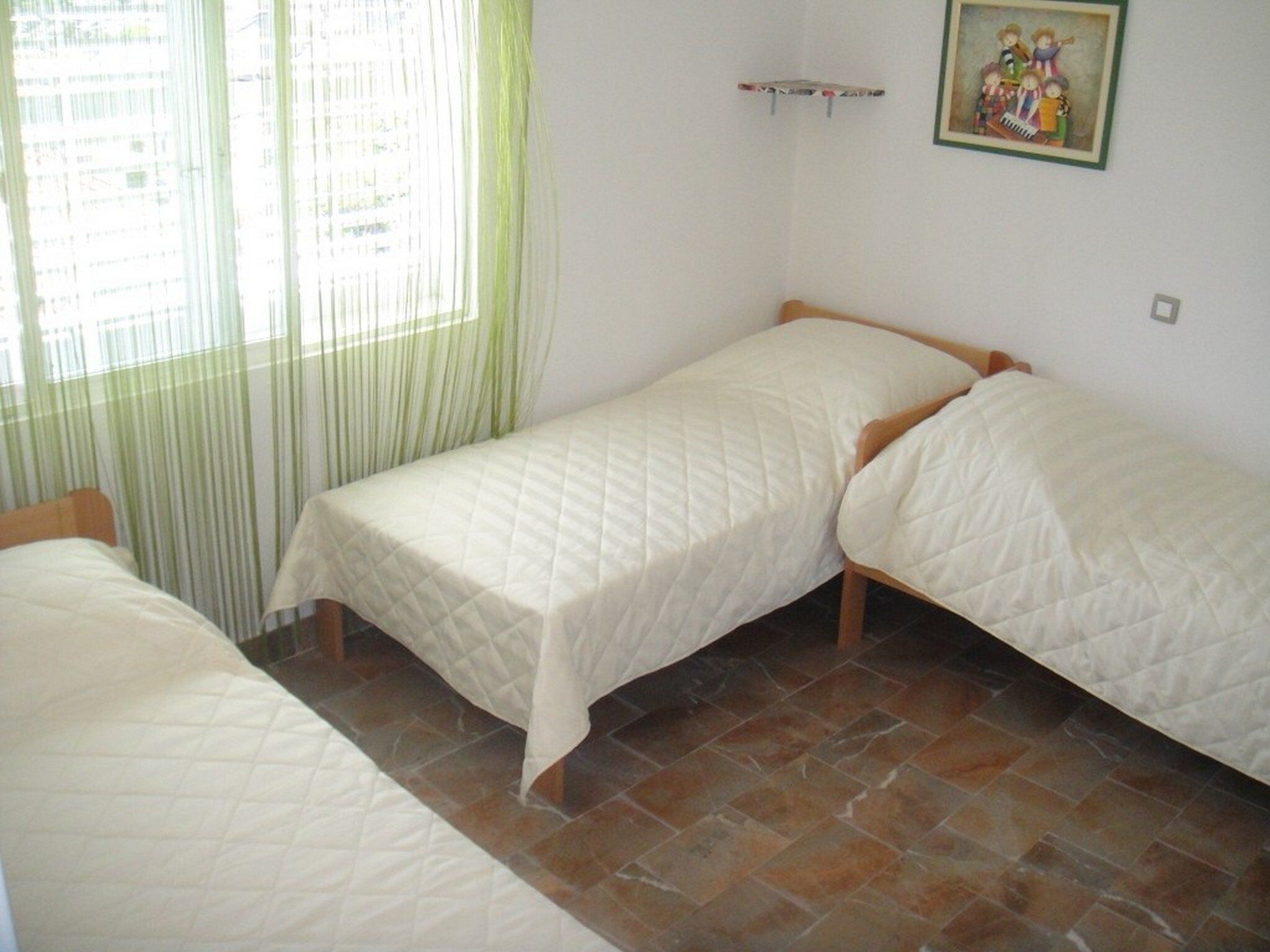 A room with two single beds, a window with green curtains, and a wall-mounted shelf with a framed picture.
