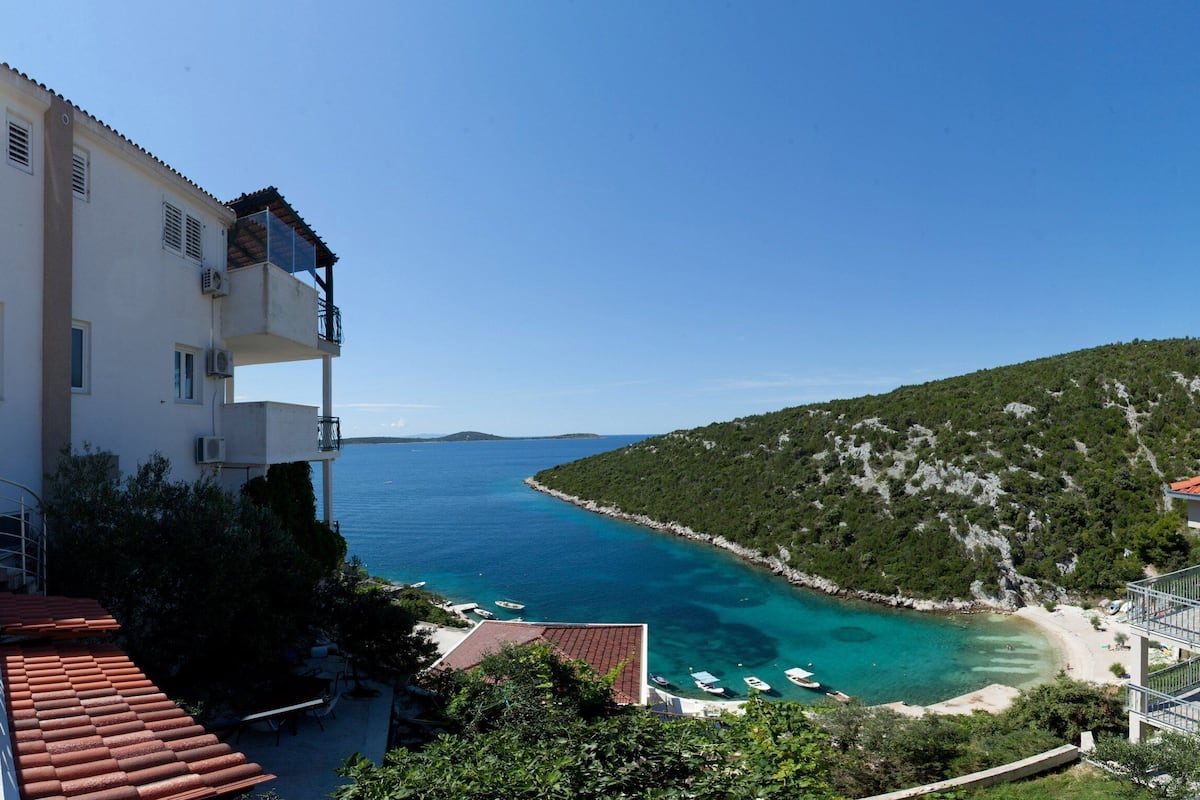 A coastal building with balconies overlooking a clear blue sea and a hilly landscape.