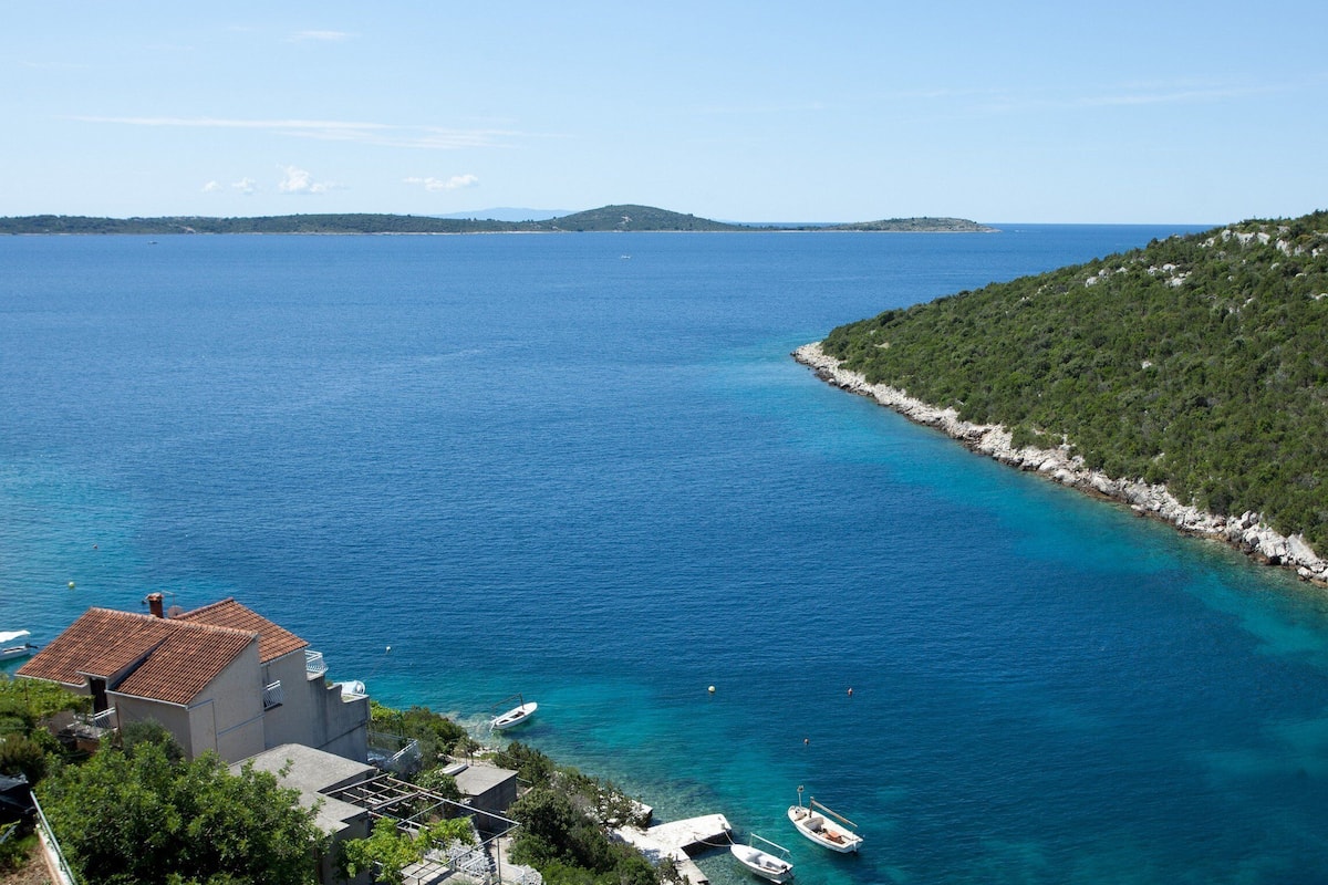 A coastal view with a clear blue sea, boats docked near the shore, and a building with a red roof.