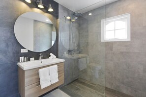 Un baño moderno con un espejo grande, un tocador de madera, una ducha con cerramiento de vidrio y una ventana que proporciona luz natural.