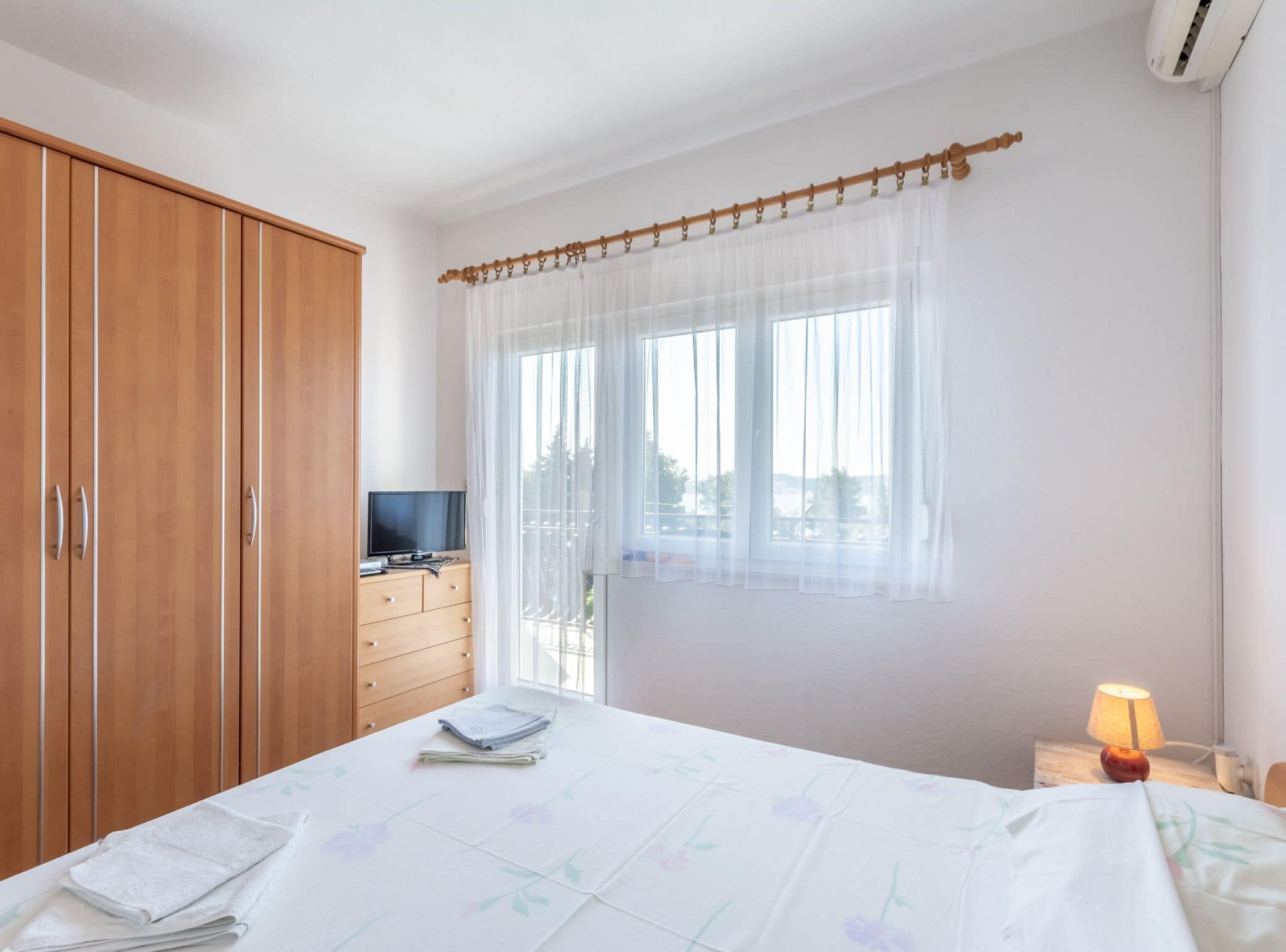 Ein Schlafzimmer mit einem Bett, einem Fernseher auf einer Holzkommode, einem Fenster mit durchsichtigen Vorhängen und einer Klimaanlage.