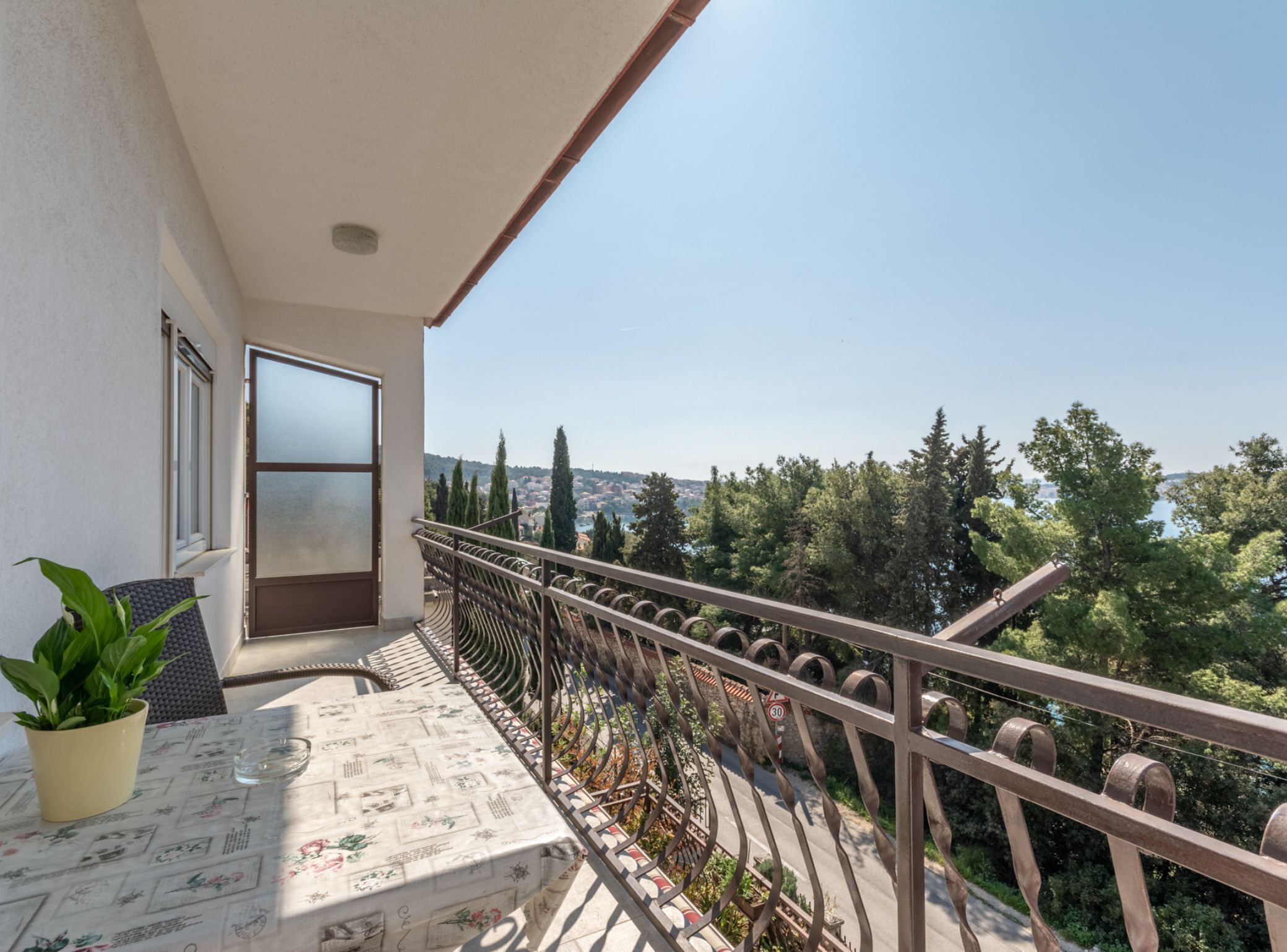 A balcony with a table, potted plant, and a view of a town and trees.