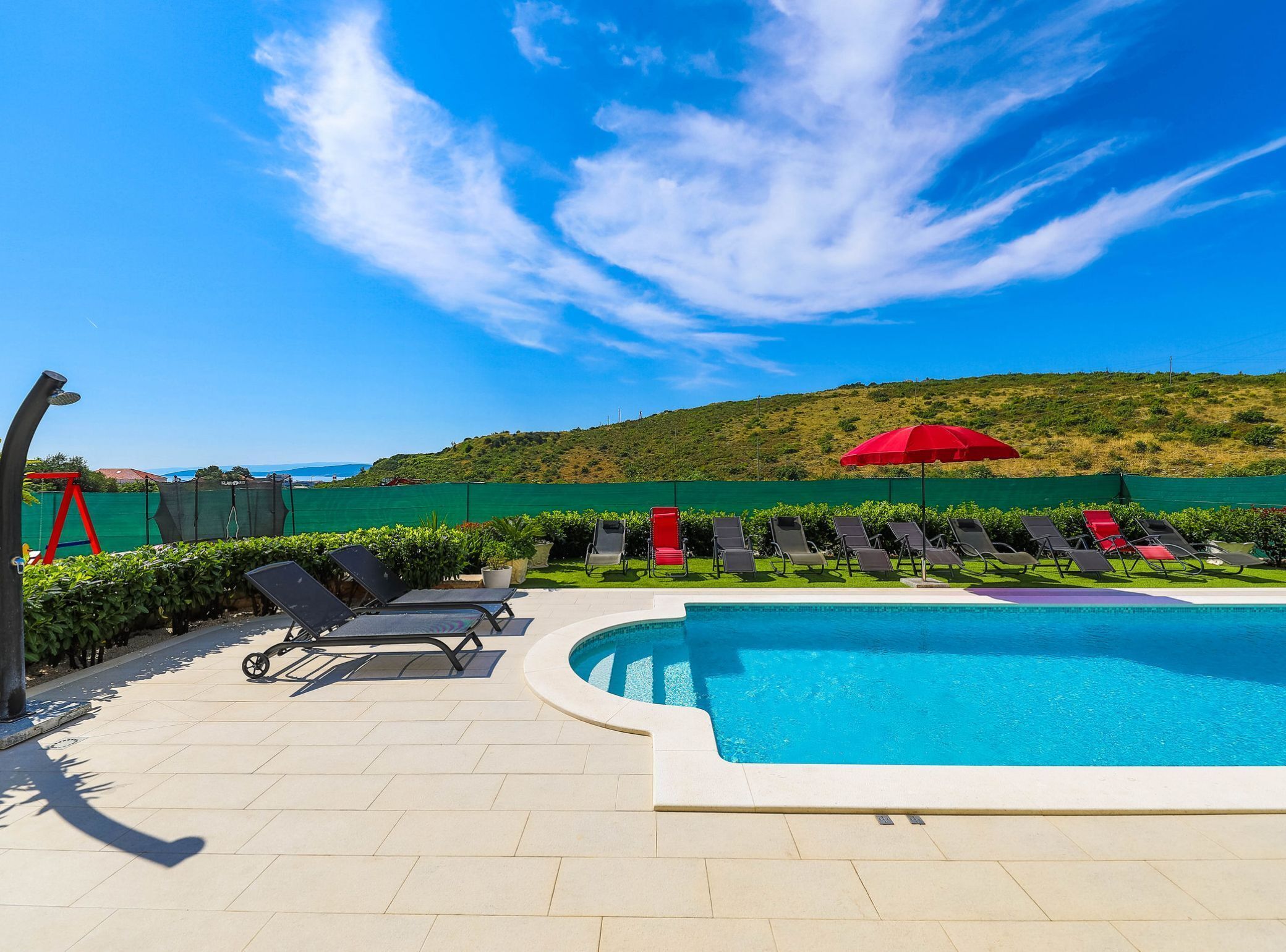 A pool area with lounge chairs, a red umbrella, and a view of a green hill and sea.