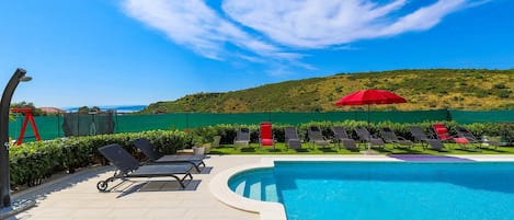 A pool area with lounge chairs, a red umbrella, and a view of a green hill and sea.